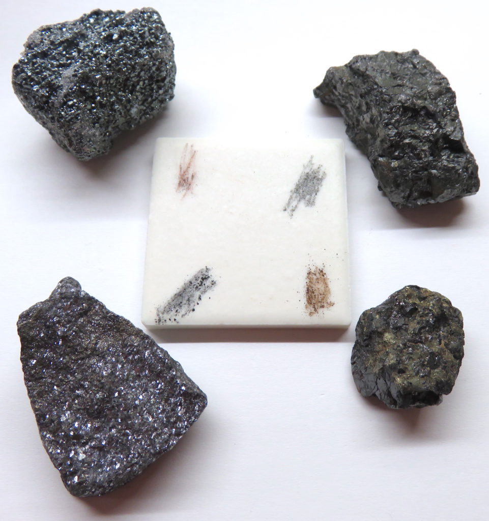 Similar dark-grey minerals with varying degrees of metallic sheen leave different colours of streaks. The minerals are from upper left clockwise: hematite, magnetite, sphalerite, and galena. _Source: Karla Panchuk (2015) CC BY 4.0_