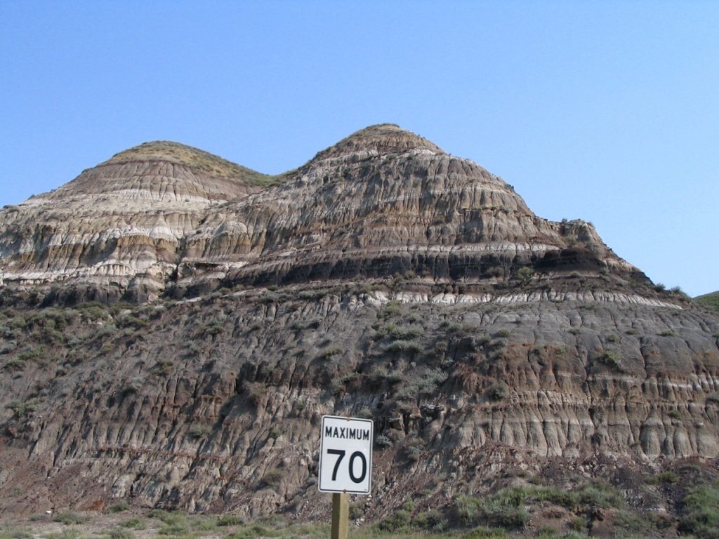 Cretaceous sedimentary rocks exposed along a road near Drumheller, Alberta, Canada. Sedimentary rocks form in layers called beds, and the planar boundaries that separate each bed are called contacts. Each bed tells a story about the conditions in which it formed. In this picture the beds are indicating that sea level repeatedly rose and fell. The black layer about halfway up the picture is a coal seam. It tells us that the environment at that time was swampy. _Source: Karla Panchuk (2008) CC BY 4.0_
