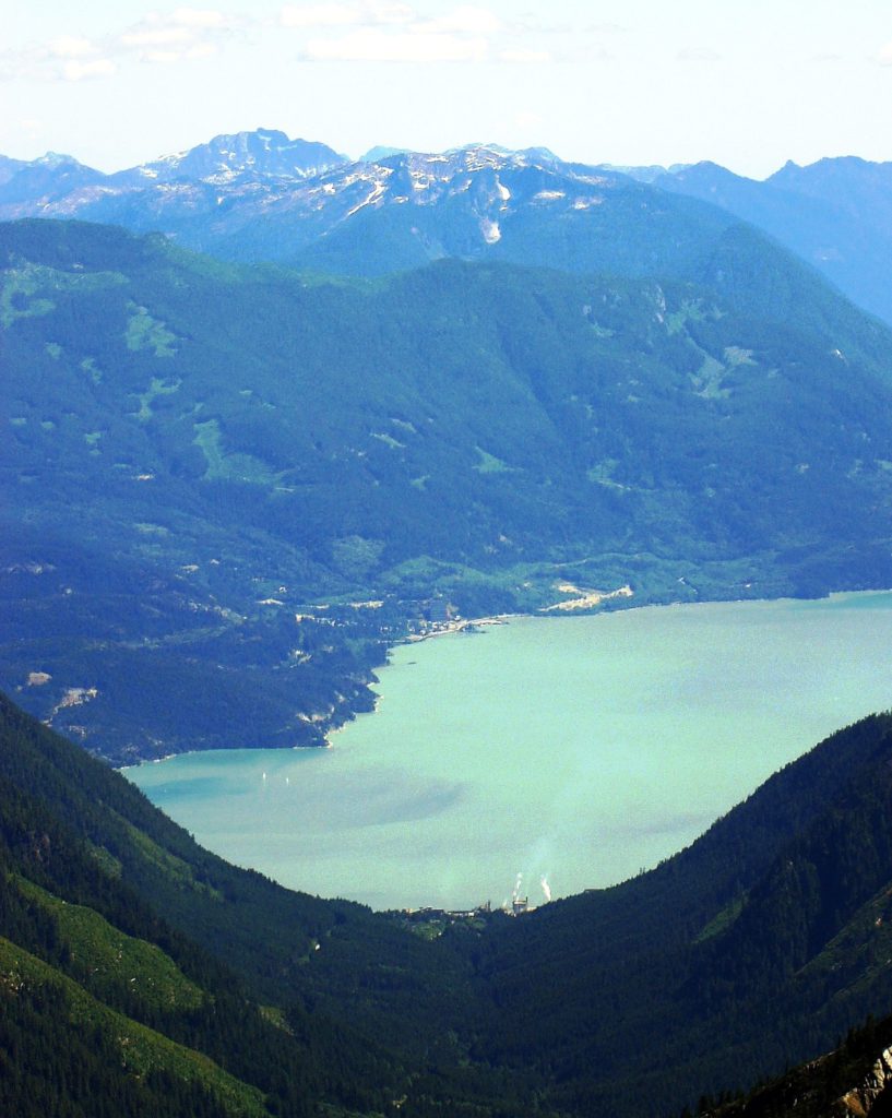 The view down the U-shaped valley of Mill Creek valley toward the U-shaped valley of Howe Sound, with the village of Britannia on the opposite side. _Source: Keefer4 (2005) CC BY-SA 2.5 [view source](https://commons.wikimedia.org/wiki/File:Woodf1a.jpg)_